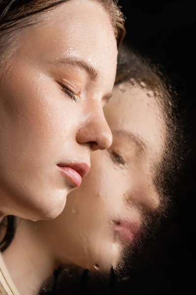 Nahaufnahme einer jungen Frau mit nasser Haut, die in der Nähe eines Spiegels auf schwarzem Hintergrund posiert — Stockfoto