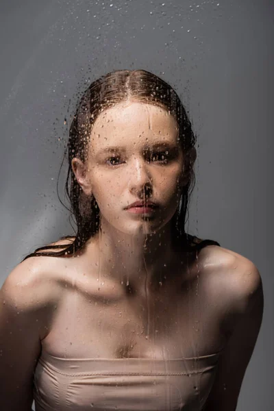 Jeune femme regardant caméra derrière verre mouillé sur fond gris — Photo de stock