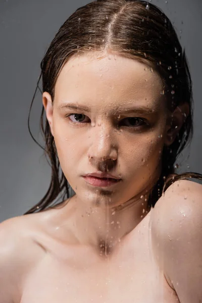Portrait of woman with naked shoulders standing behind wet glass on grey background — Stock Photo