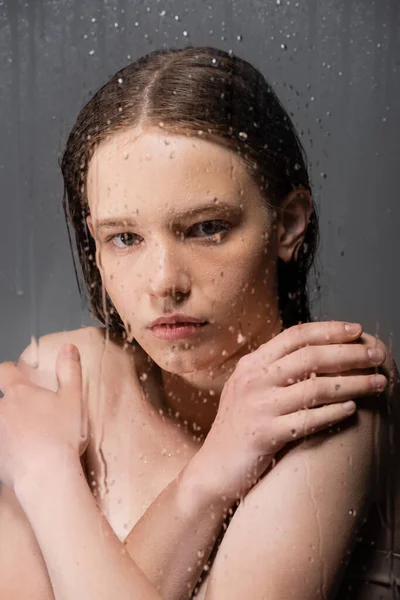 Portrait de jeune femme touchant les épaules derrière un verre mouillé sur fond gris — Photo de stock