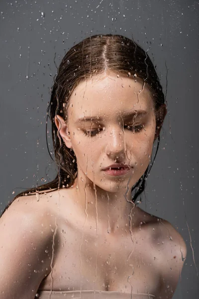 Светловолосая модель с голыми плечами, стоящая за мокрым стеклом на сером фоне — стоковое фото
