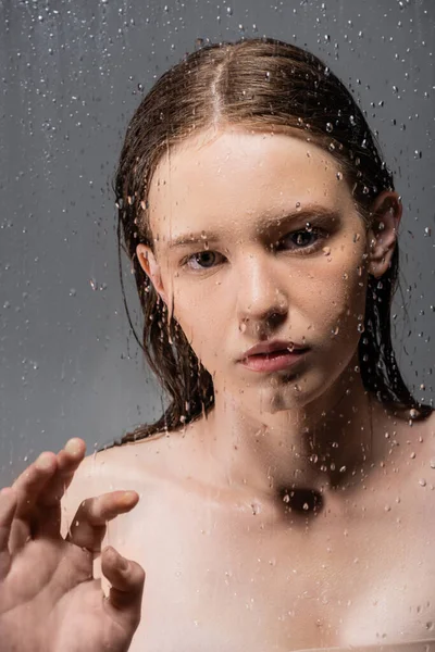 Young model with naked shoulders touching wet glass on grey background — Stock Photo