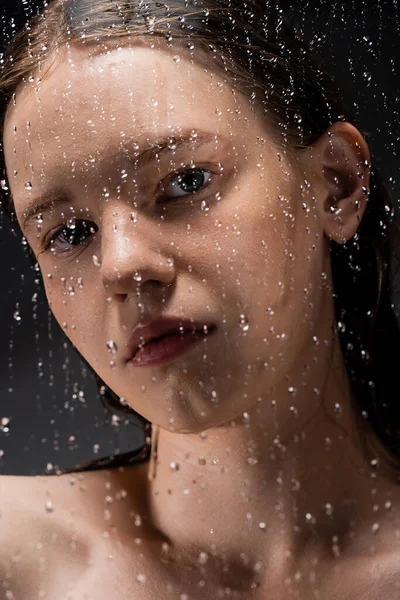 Portrait of young woman looking at camera through wet glass on grey background — Stock Photo