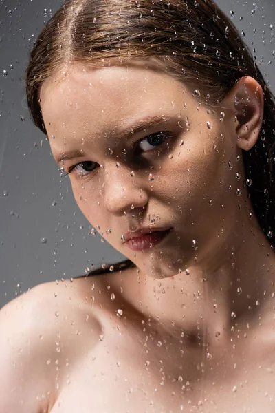 Schönhaariges Model mit nackter Schulter, das hinter nassem Glas auf grauem Hintergrund in die Kamera blickt — Stockfoto