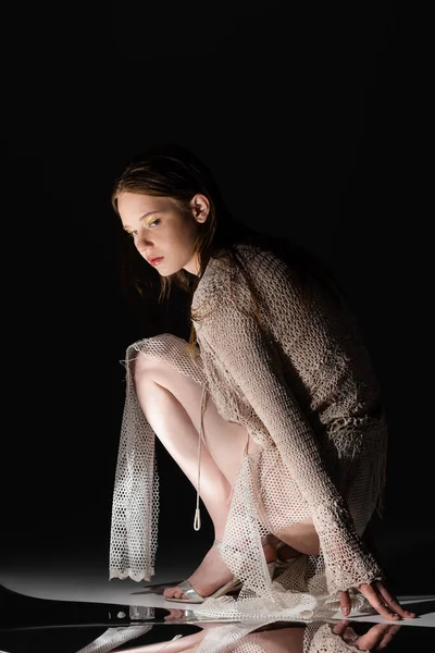 Elegante giovane donna in gonna a rete e cardigan lavorato a maglia posa vicino superficie riflettente su sfondo nero — Foto stock