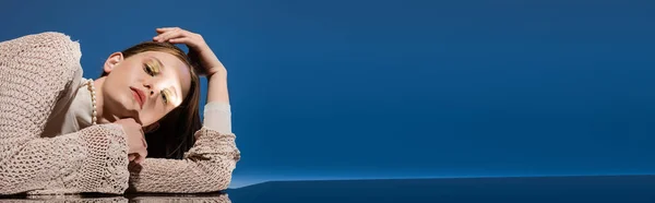 Модель из жемчужного ожерелья и вязаного кардигана лежащая на отражающей поверхности на градиентно-синем фоне, баннер — стоковое фото
