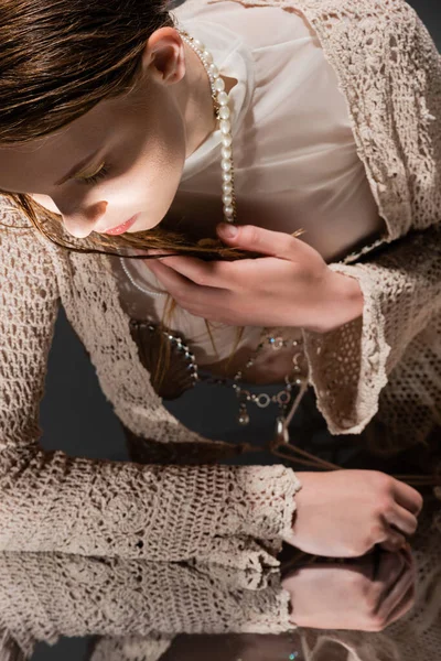 Женщина в вязаном кардигане и жемчужном ожерелье, глядя на зеркало на сером фоне — стоковое фото