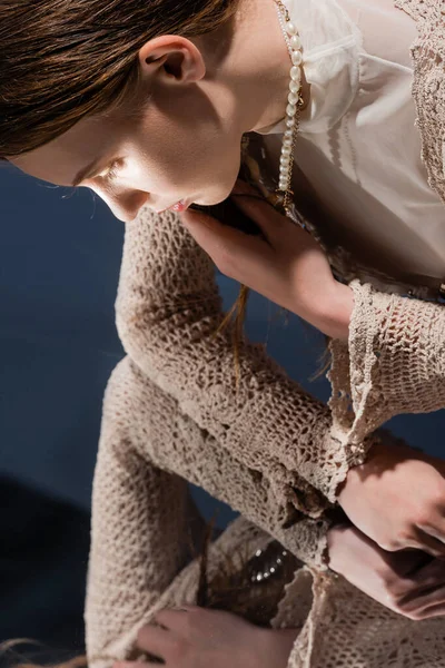 Vista laterale della donna in maglia cardigan e collana di perle guardando specchio su sfondo blu — Foto stock