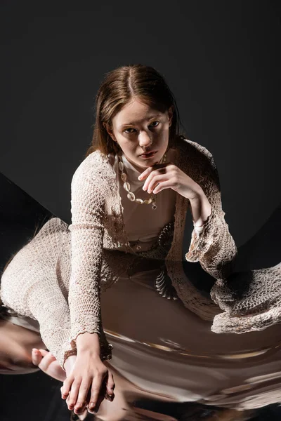 Stilvolle junge Frau in Muschelhalskette und Strickjacke berührt reflektierende Oberfläche auf grauem Hintergrund — Stockfoto