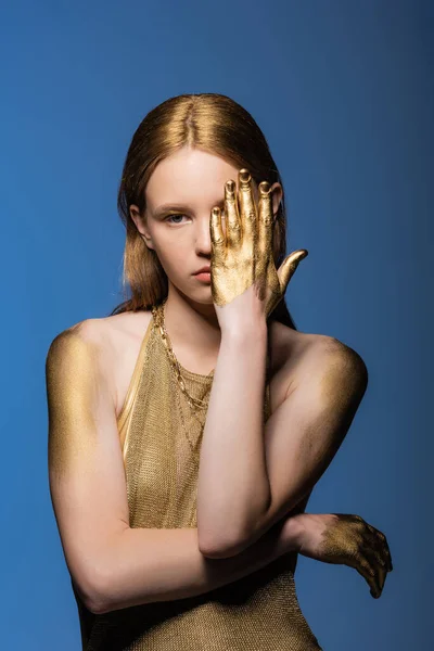 Mujer de pelo rubio en pintura dorada en las manos que cubre el ojo aislado en azul - foto de stock