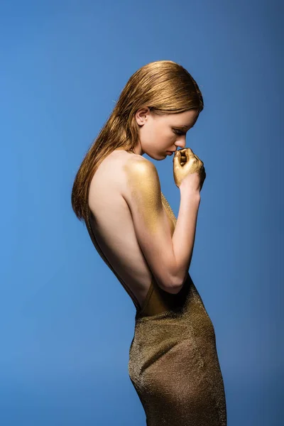 Seitenansicht der jungen Frau in goldenem Kleid und Farbe berühren Lippen isoliert auf blau — Stockfoto