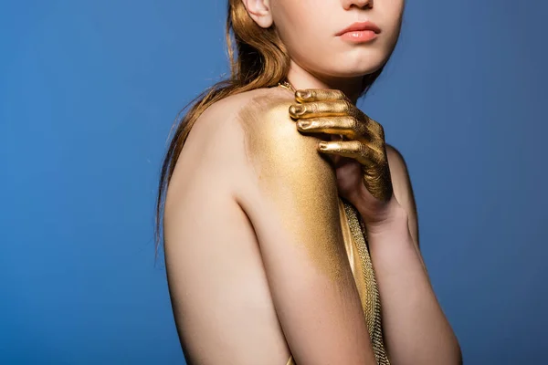 Обрезанный вид молодой женщины с золотой краской на руке касаясь плеча изолированы на синий — стоковое фото