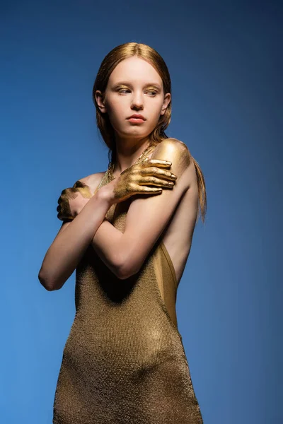 Modelo de pelo limpio en vestido y pintura dorada en las manos tocando hombros aislados en azul - foto de stock