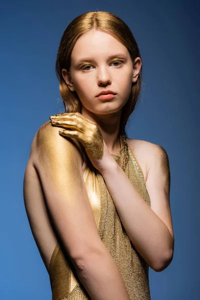 Porträt eines jungen Models in goldenem Kleid und Farbe, das isoliert auf blauem Grund in die Kamera blickt — Stockfoto