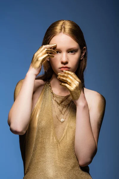 Modelo elegante en vestido y manos en pintura dorada tocando la cara aislada en azul - foto de stock