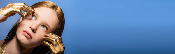 Портрет молодой женщины с золотой краской на руках и волосы касаясь лица изолированы на синий, баннер — стоковое фото