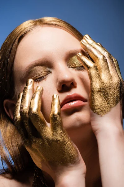 Portrait of young woman with makeup and golden paint on hands touching face isolated on blue — Stock Photo