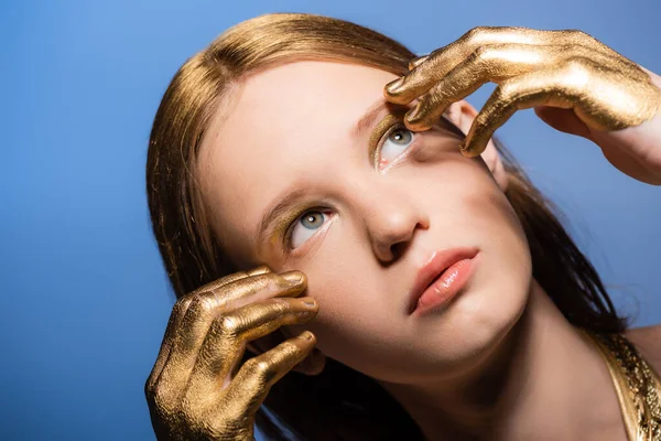Mujer joven y bonita con pintura dorada en las manos tocando la cara y mirando hacia otro lado aislado en azul - foto de stock