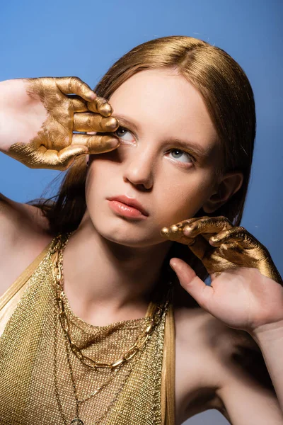 Junge Frau mit goldener Farbe an den Händen, die wegschaut, während sie isoliert auf blau posiert — Stockfoto