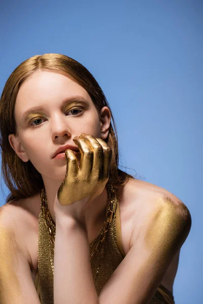 Porträt einer Frau mit goldener Farbe auf Schultern und Hand, die isoliert auf blauem Grund in die Kamera blickt — Stockfoto