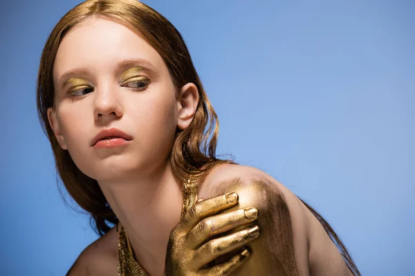 Jeune modèle aux cheveux blonds avec peinture dorée à la main touchant épaule isolée sur bleu — Photo de stock