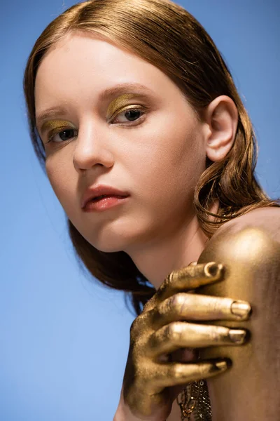 Porträt einer jungen Frau mit goldenem Make-up und Farbe auf der Hand, die isoliert auf blauem Grund in die Kamera blickt — Stockfoto