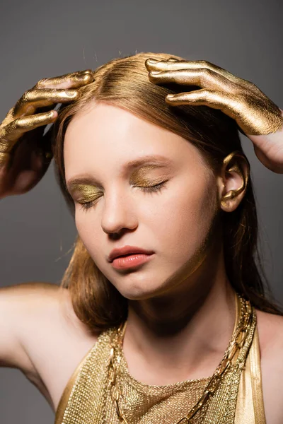 Modelo joven con maquillaje y manos en pintura dorada tocando el cabello aislado en gris - foto de stock