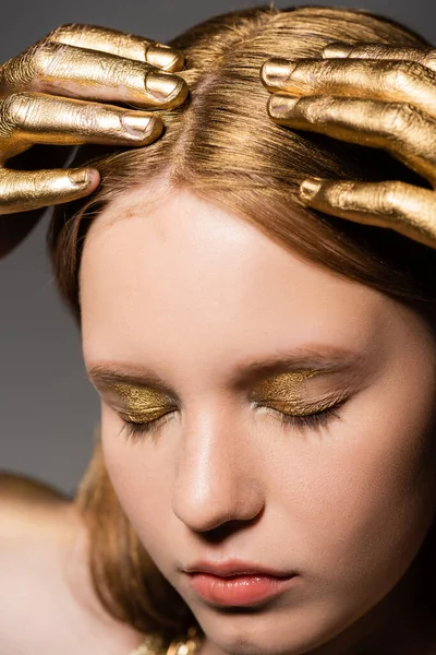 Portrait of young woman with golden makeup and paint on hands touching hair and closing eyes isolated on grey — Stock Photo