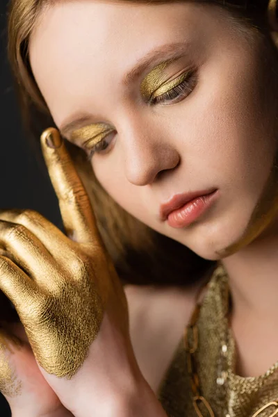 Retrato de mujer joven con maquillaje dorado y pintura en la mano tocando la ceja aislada en gris - foto de stock