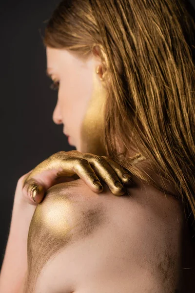 Боковой вид зарытой женщины с золотой краской на руке, касающейся плеча, изолированного на сером — стоковое фото