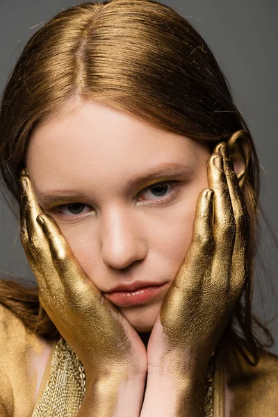 Portrait of young woman with golden paint on hands touching face isolated on grey — Stock Photo