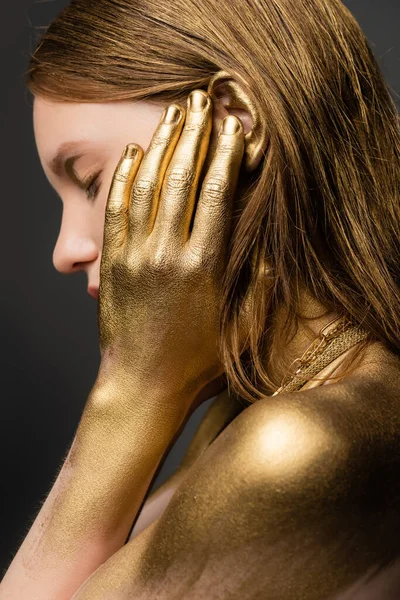Вид збоку молодої жінки в золотій фарбі, що торкається вуха, ізольовані на сірому — стокове фото