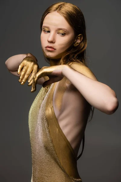 Mujer joven con pintura dorada en la cara y las manos mirando hacia otro lado aislado en gris - foto de stock