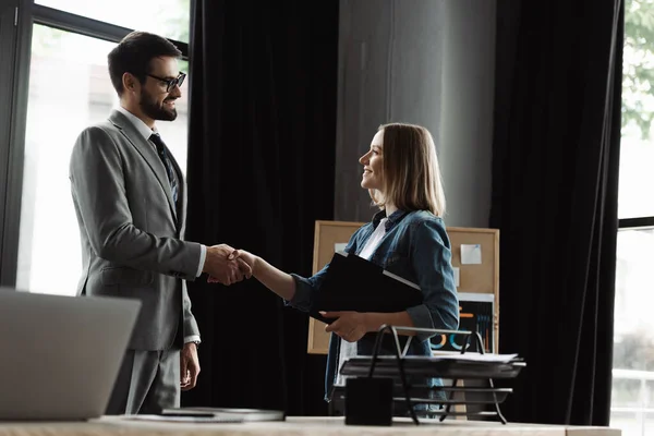 Mujer sonriente sosteniendo curriculum vitae y estrechando la mano con el hombre de negocios durante la entrevista de trabajo en la oficina - foto de stock