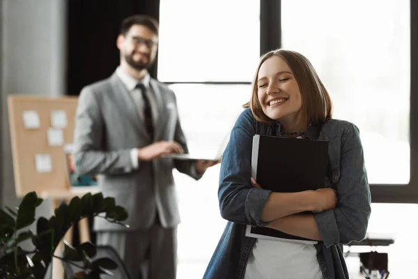 Позитивна жінка тримає паперову теку з резюме біля розмитого бізнесмена в офісі — стокове фото
