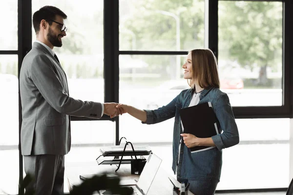 Side view of smiling businessman shaking hand of young woman with resume in office — Stock Photo