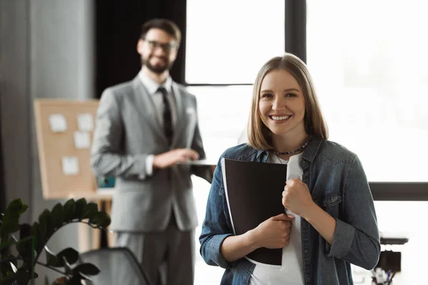 Cheerful woman with resume showing yes gesture near blurred businessman in office — Stock Photo