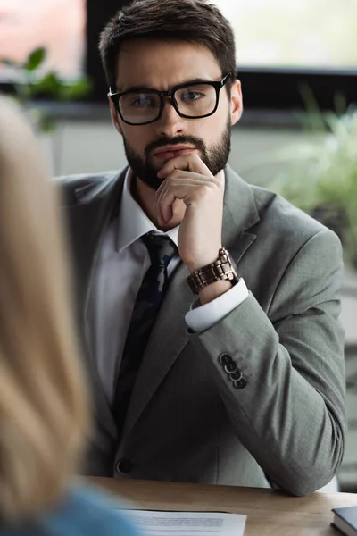Hombre de negocios concentrado en traje mirando a la mujer borrosa durante la entrevista de trabajo en la oficina - foto de stock