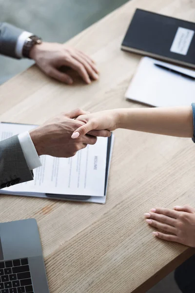 Cropped view of businessman and job seeker shaking hands near blurred resume in office — Stock Photo