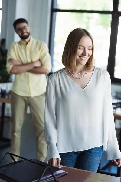 Smiling woman standing near working table near blurred colleague during internship in office — Stock Photo