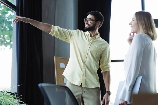 Hombre de negocios sonriente señalando con el dedo a la ventana cerca de pasante con portátil en la oficina - foto de stock