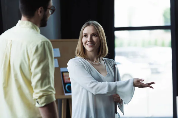 Smiling intern with laptop pointing with hand near blurred businessman in office — Stock Photo