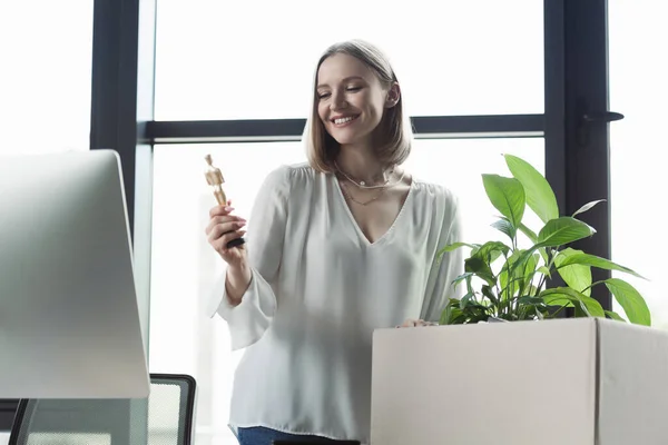 Fröhlicher Praktikant hält Puppe neben Karton mit Pflanze und Computer im Büro — Stockfoto