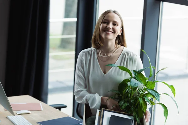 Happy intern looking at camera near cardboard box with stuff and computer in office — Stock Photo