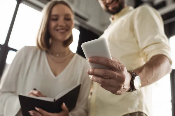 Low angle view of smartphone in hand of blurred businessman near intern with notebook in office — Stock Photo