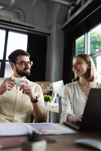 Estagiário alegre olhando para homem de negócios com xícara de café perto de laptop e papéis no escritório — Fotografia de Stock