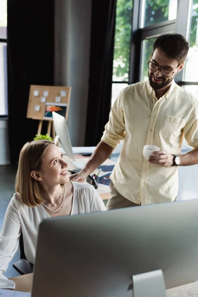 Lächelnder Manager mit Kaffeetasse neben neuem Mitarbeiter und Computer im Büro — Stockfoto