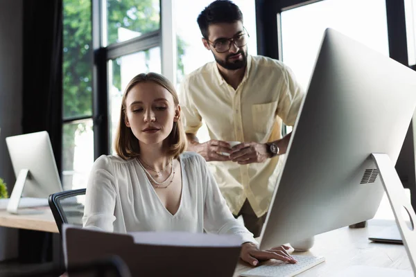 Pasante mirando los papeles y el uso de la computadora cerca borrosa hombre de negocios con café - foto de stock