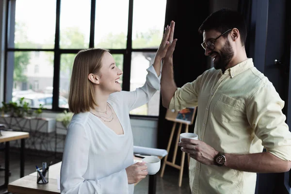 Side view of intern and manager with coffee cups giving high five in office — Stock Photo