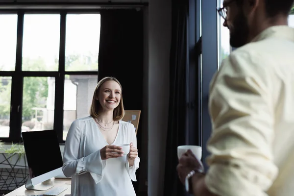 Cheerful new worker holding cup of coffee near blurred businessman in office — Stock Photo